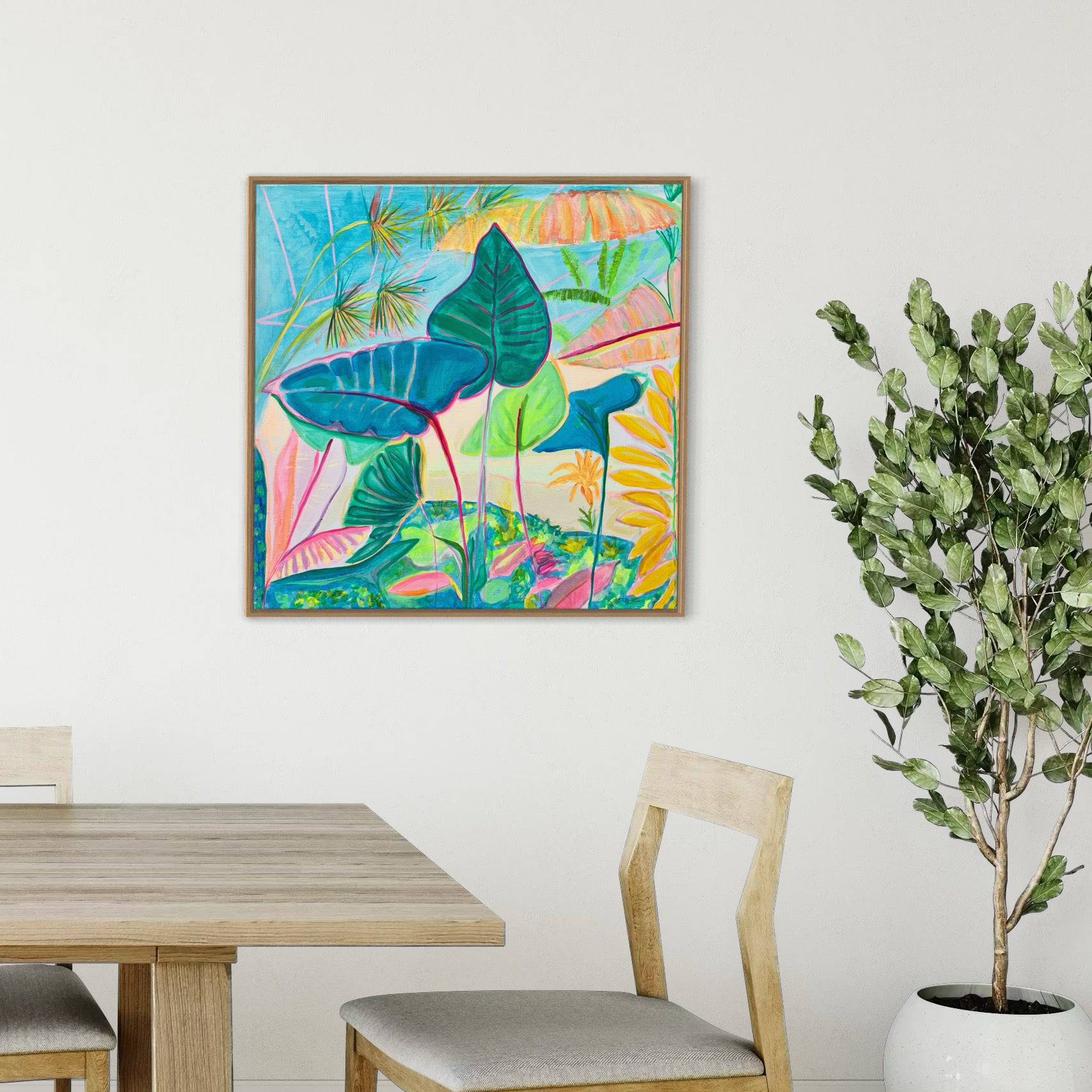 Colorful botanical painting by Kathryn Sillince with blue and green leaves in a simple wooden frame on a white wall above a wooden dining table and chair, next to a potted green plant