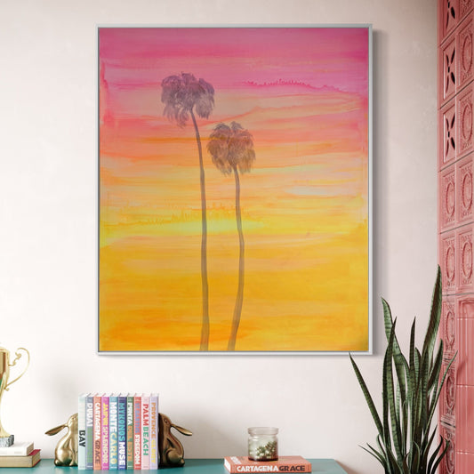 Framed artwork of palm trees against a sunset sky on a wall above books and decor items.